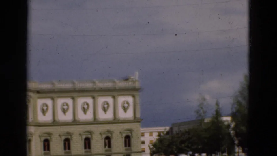 1975 Portugal 8mm frame showing the upper part of a classical facade, blue sky, nearby trees, and a distant pale building.