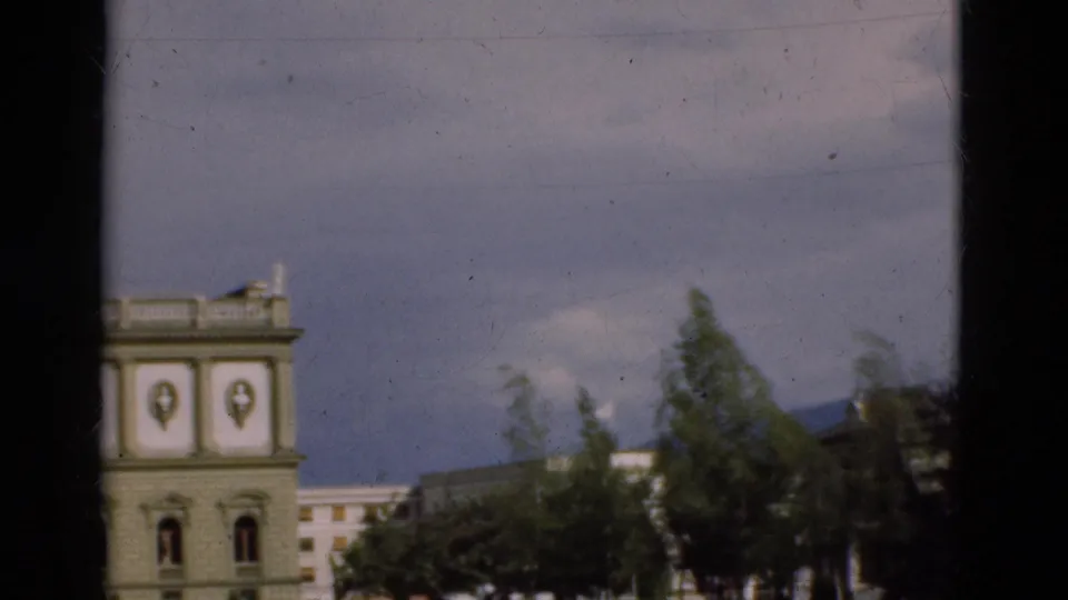 1975 Portugal home-movie frame with part of a pale classical facade, trees in the foreground, and low buildings beyond under blue sky.