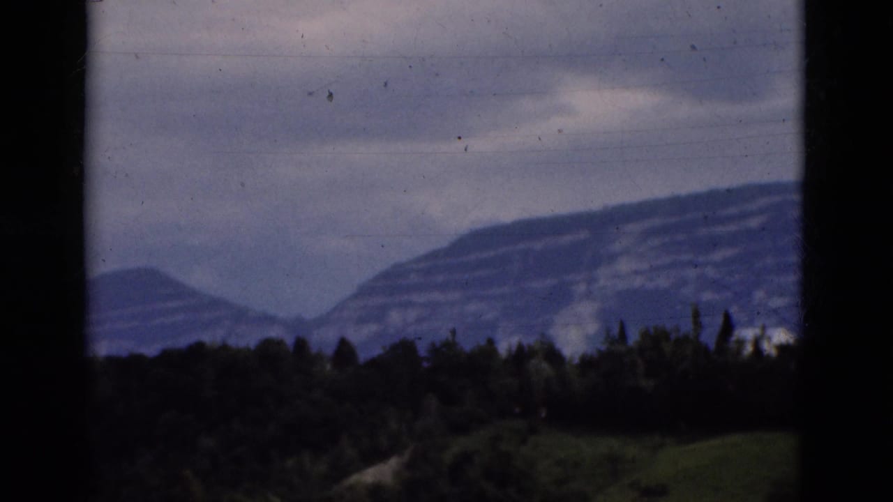 Layered mountains and dark tree line under a cloudy sky, partly framed by black edges.