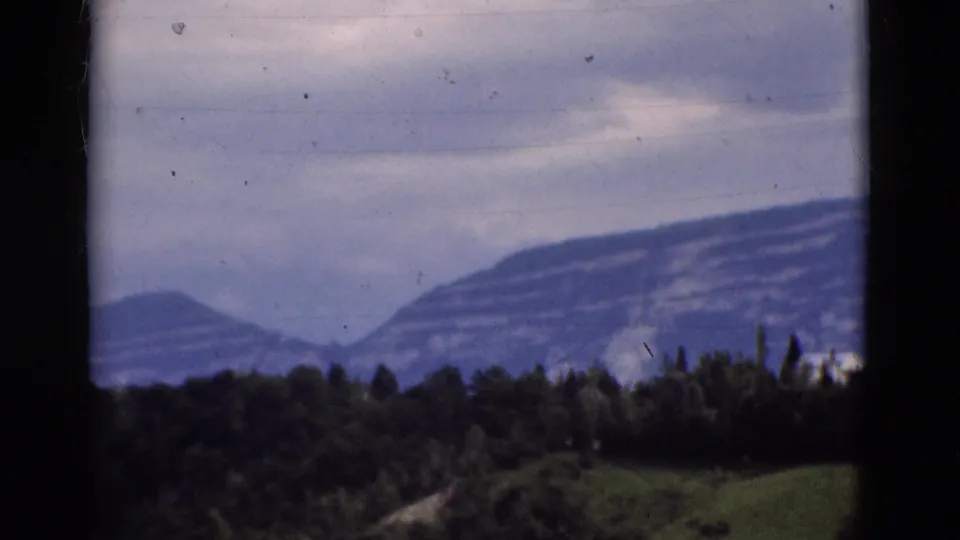 1975 Portugal landscape frame with a broad mountain slope, dark forested foreground, and cloudy sky on soft 8mm film.