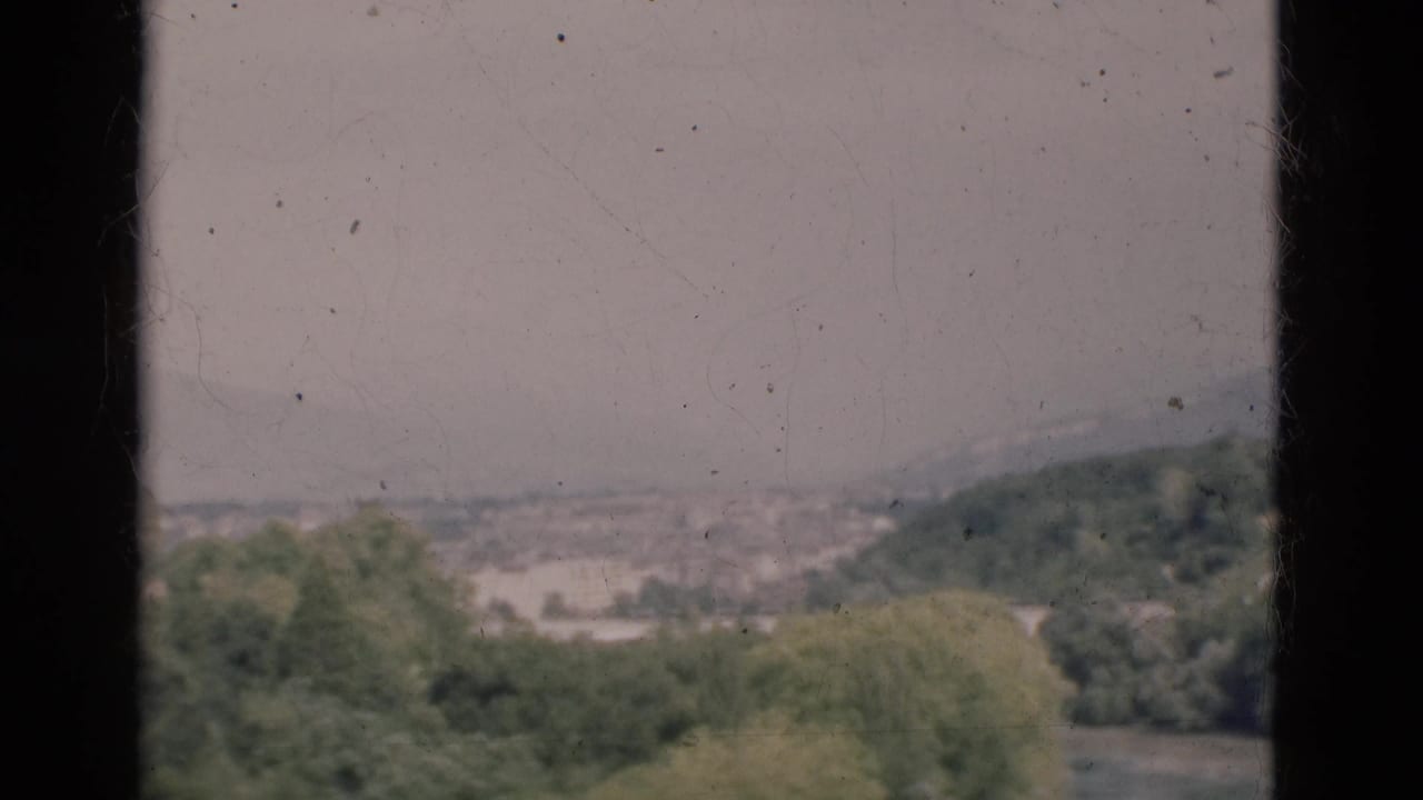 1975 Portugal home-movie frame with a hazy hillside city view seen through dark vertical edges and soft 8mm grain.