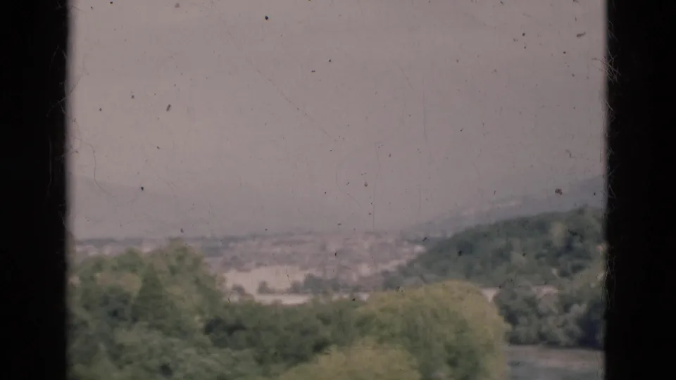 1975 Portugal home-movie frame with a hazy hillside city view seen through dark vertical edges and soft 8mm grain.
