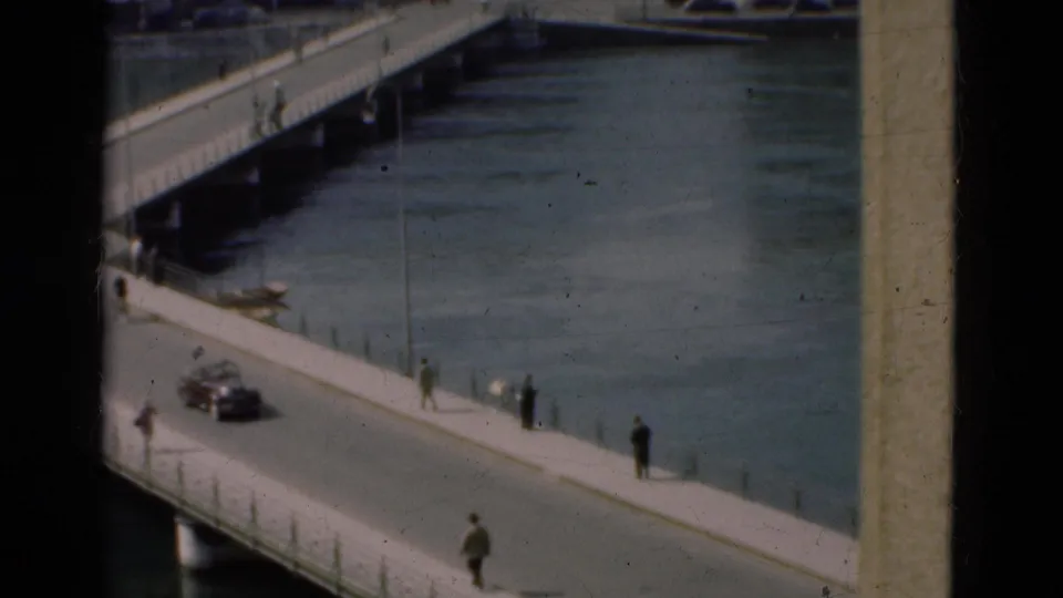 1975 Portugal frame showing a road bridge over water, with pedestrians on the sidewalk and a dark car crossing.