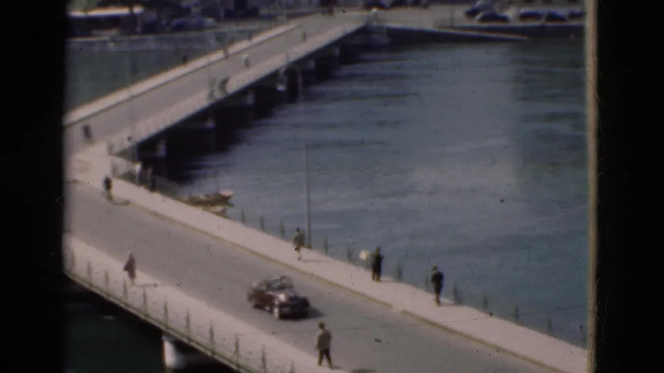 8mm home-movie frame from Portugal in 1975 showing a dark car on the bridge and several pedestrians near the river railing.