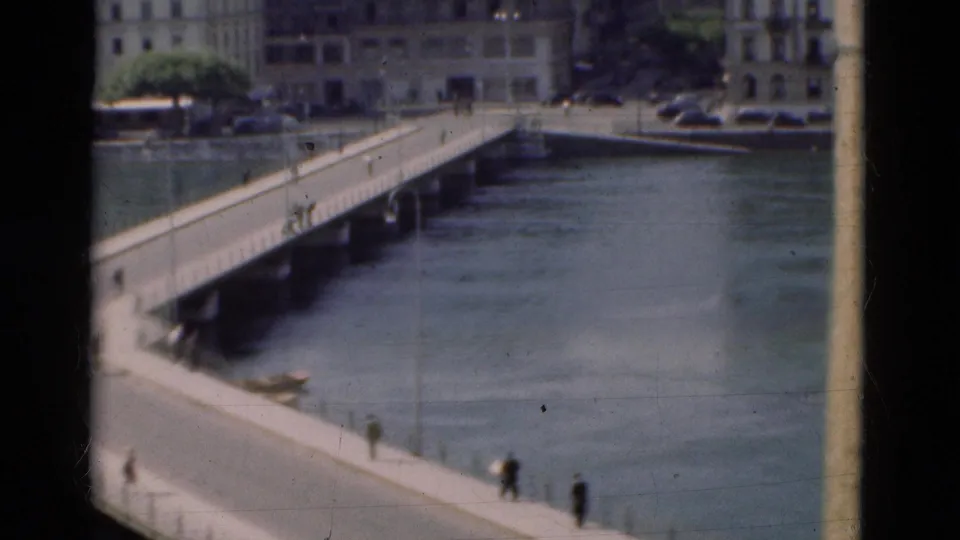 Wider 1975 Portugal bridge view with pedestrians over the river, waterfront buildings, parked cars, and soft 8mm film texture.