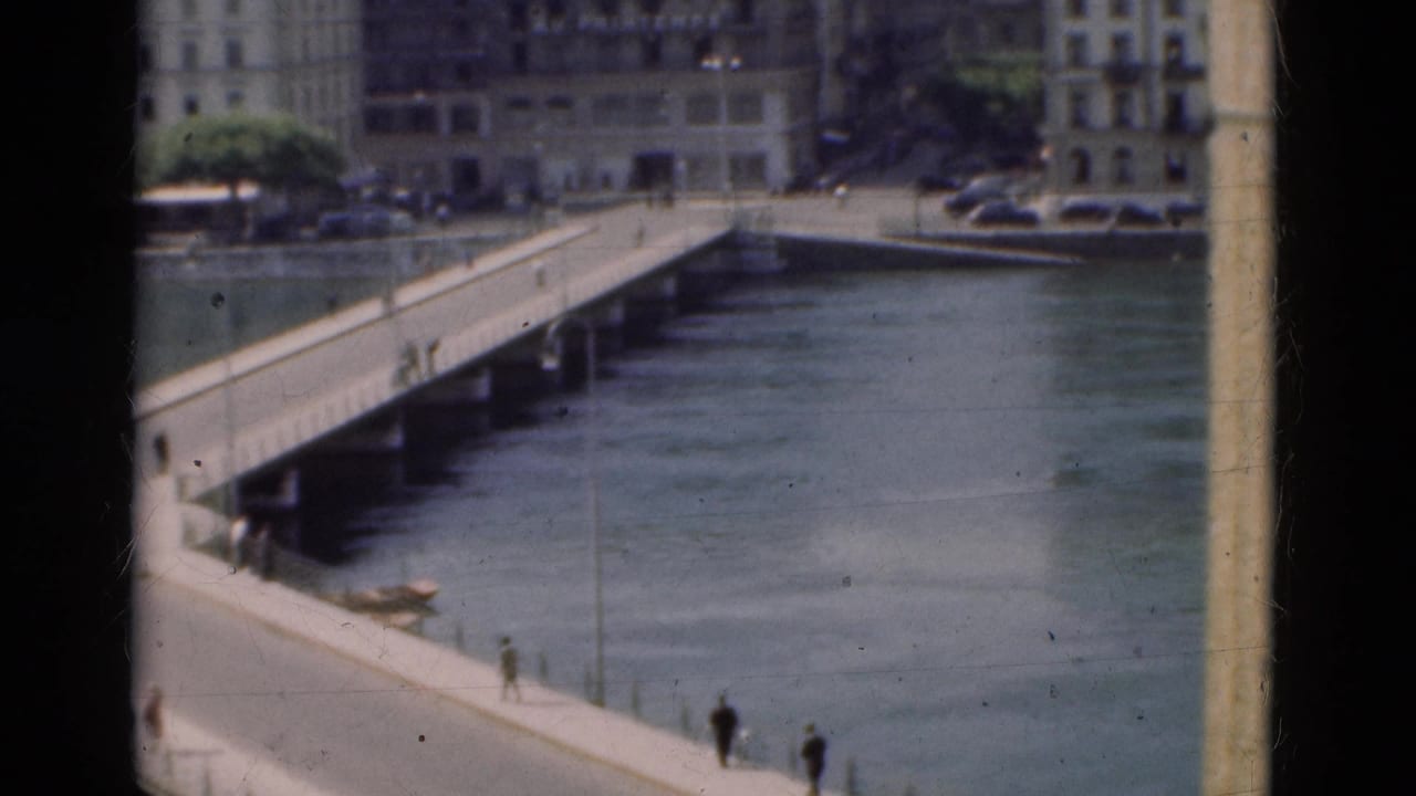 Pedestrians cross a long bridge over calm water with buildings on the far bank.