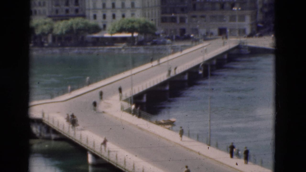1975 Portugal frame of pedestrians scattered along a bridge over water, with urban buildings and trees beyond.