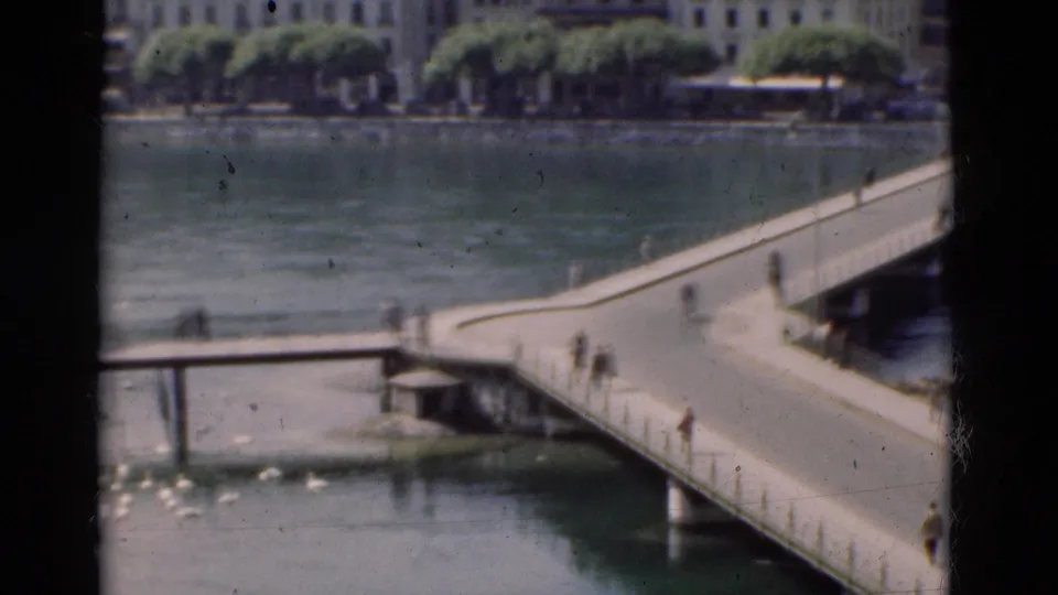 1975 Portugal frame showing pedestrians on a bridge over calm water, with waterfront buildings and trees in the background.