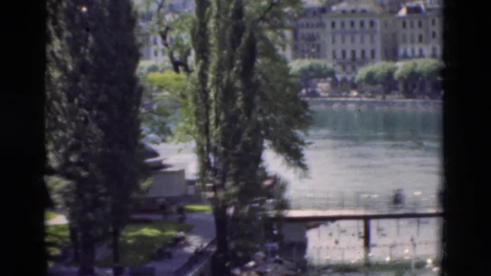 1975 Portugal home-movie frame looking past tall trees to a lake, shoreline walkway, water birds, and distant European buildings.