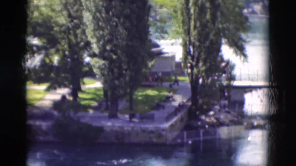 1975 Portugal 8mm frame of a tree-lined riverbank with grass, stone edges, paths, water, and small distant people.
