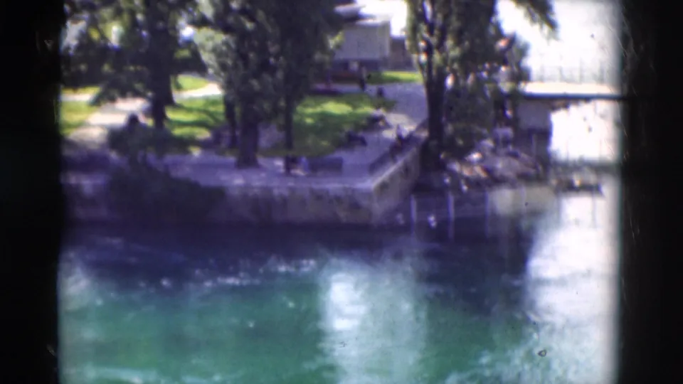 1975 Portugal home-movie frame showing a stone river corner, tall trees, grass, and bright reflected water.