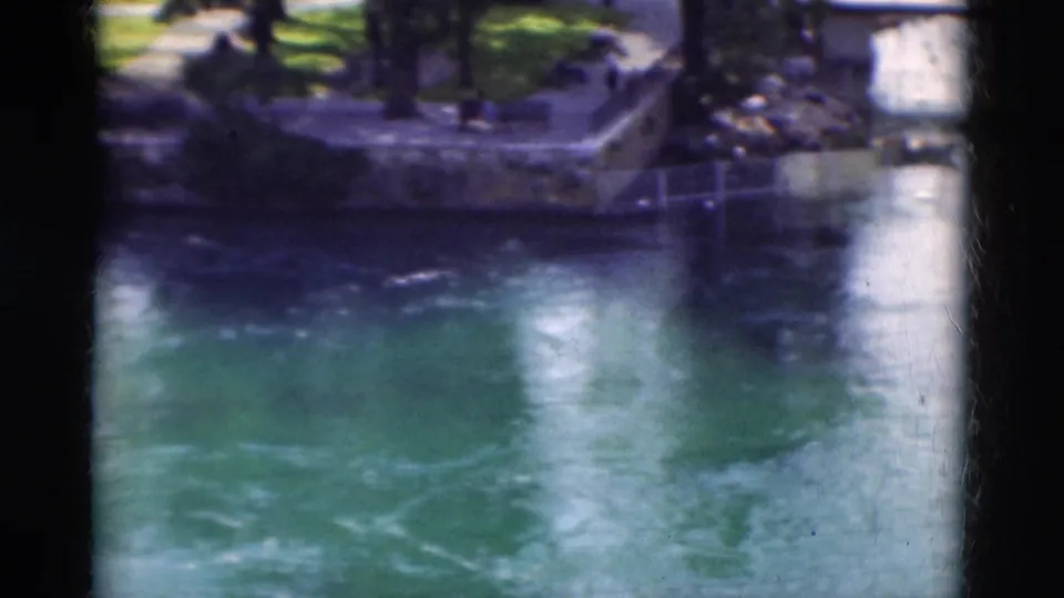 1975 Portugal 8mm frame with blue-green water in the foreground and a shaded stone riverbank with trees and distant people.