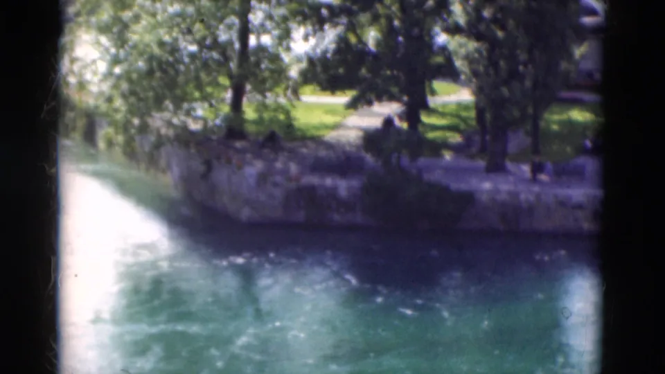 1975 Portugal home-movie frame showing a sunlit river, stone bank, trees, grass, and small distant park figures.