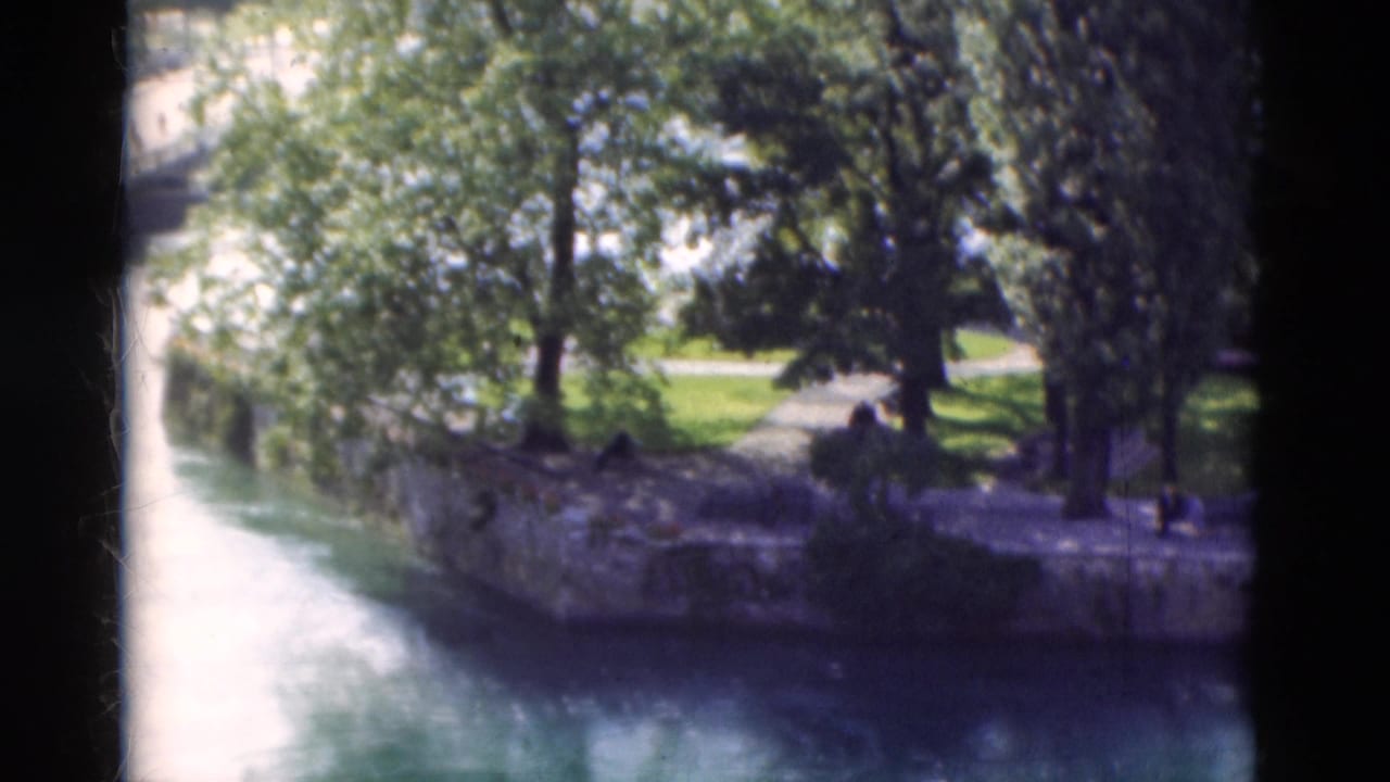 1975 Portugal 8mm frame with a curving waterway, stone wall, leafy trees, lawn, and small figures in shade.
