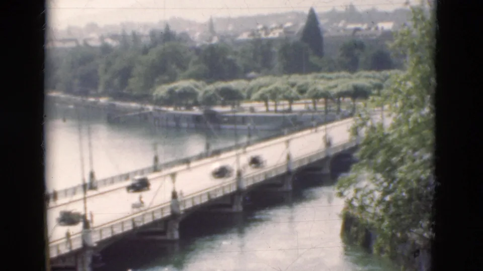 1975 Portugal 8mm frame with vintage cars on a low river bridge, landscaped banks, trees, distant buildings, and small pedestrians.
