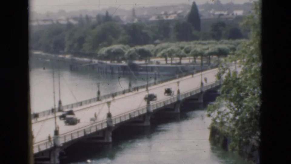 1975 Portugal home-movie view of cars crossing a river bridge, with lampposts, railings, tree-lined banks, and hazy buildings beyond.