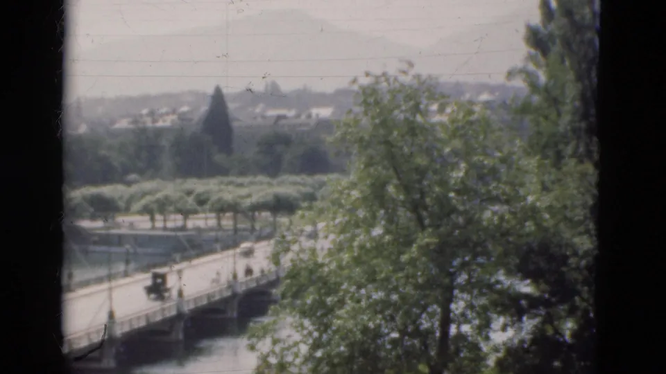Soft 8mm frame of a Portugal river bridge in 1975, showing cars, lamps, railings, trees, water, and distant landscape.