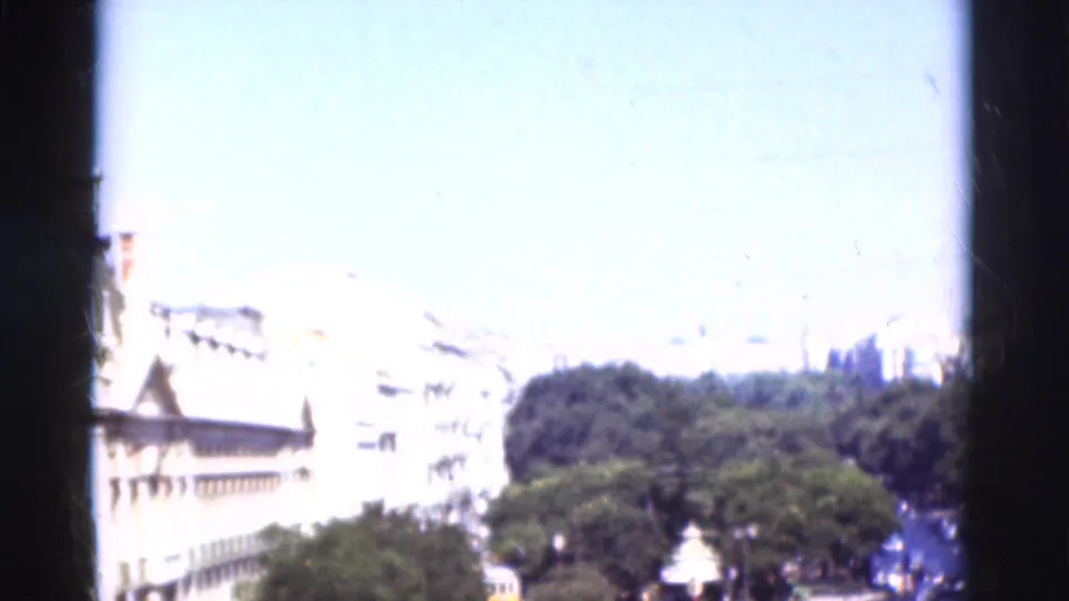 Brief 1975 Portugal urban exterior with white multi-story buildings, trees, bright sky, and heavy dark film-edge obstruction.