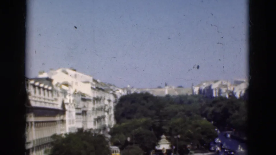 1975 Portugal 8mm frame showing an elevated city view with pale buildings, dense trees, a roadway, and clear sky.