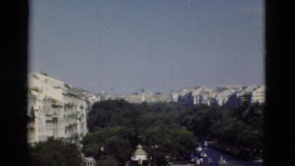 1975 Portugal home-movie frame with white urban facades, dark green trees, and a road receding through the city.