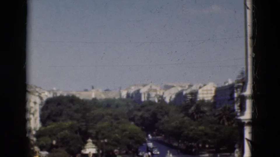 1975 Portugal 8mm frame showing rooftops, street trees, a road with vehicles, and a vertical foreground pole near the right edge.