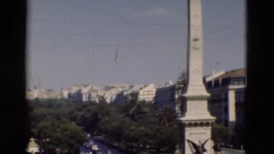 1975 Portugal frame with an obelisk-like monument entering beside tree-lined streets and light-colored city buildings.