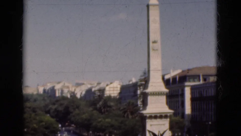 1975 Portugal 8mm frame showing a white obelisk-like monument, trees, and urban buildings from a high visitor viewpoint.