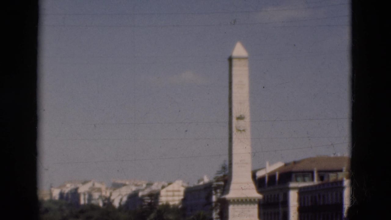 Tall white monument beside a large building with treetops and pale buildings in the background.
