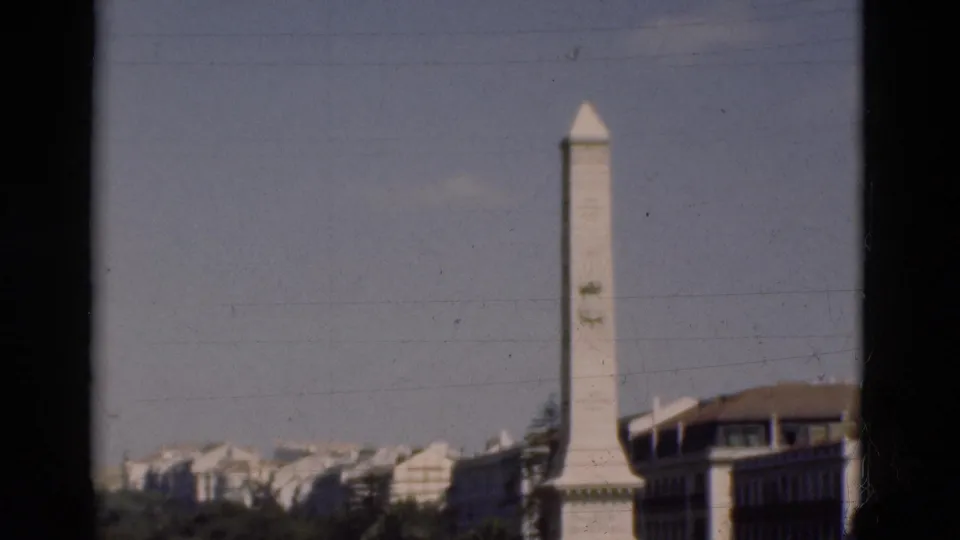 1975 Portugal frame with a tall white monument, nearby urban building, treetops, and hazy city architecture.