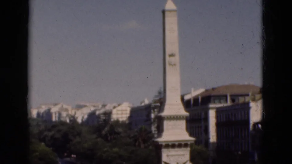 1975 Portugal home-movie frame showing the white monument centered near trees, buildings, and visible film gate edges.