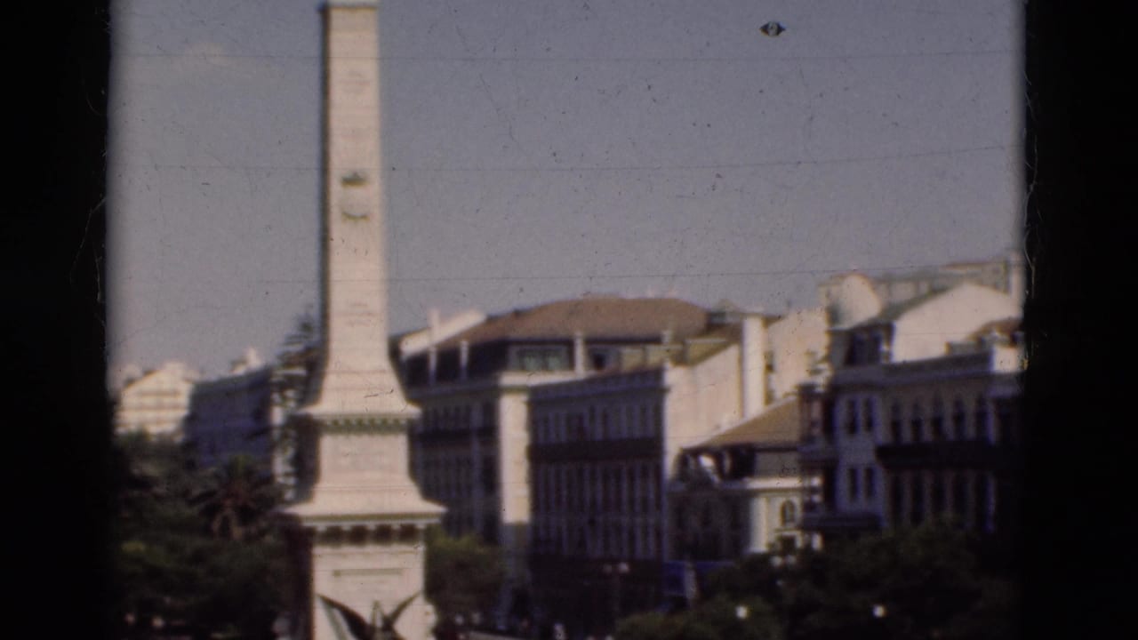 1975 Portugal home-movie frame showing a pale obelisk monument, nearby buildings, trees, and part of a roundabout from above.