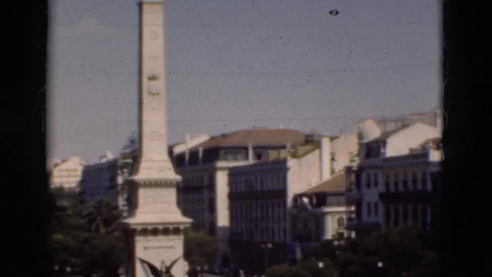 1975 Portugal home-movie frame showing a pale obelisk monument, nearby buildings, trees, and part of a roundabout from above.
