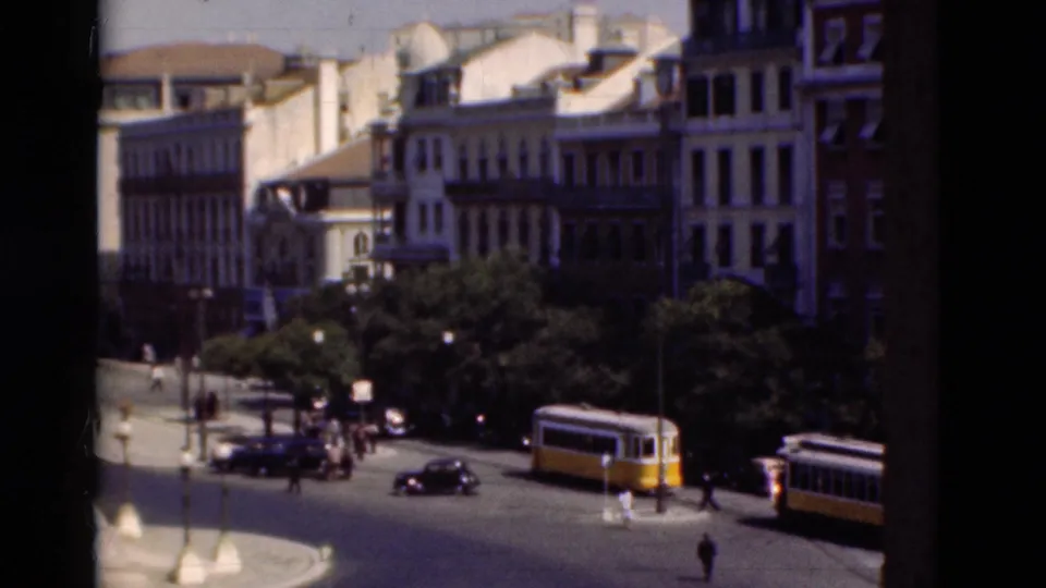 1975 Portugal street scene with yellow-and-white trams, vintage cars, pedestrians, trees, and urban buildings around a roundabout.