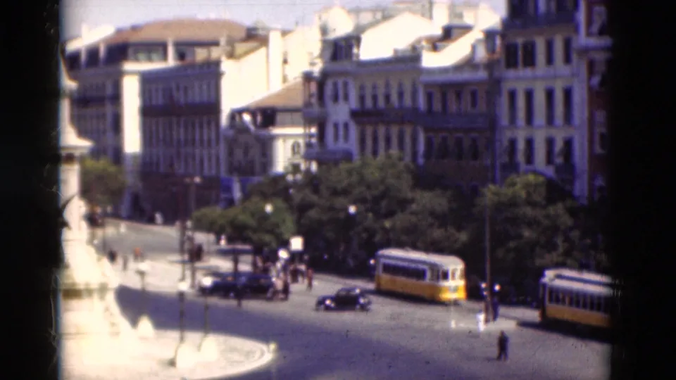 1975 Portugal elevated home-movie view of a yellow tram, cars, pedestrians, trees, and bright stone paving at a city roundabout.