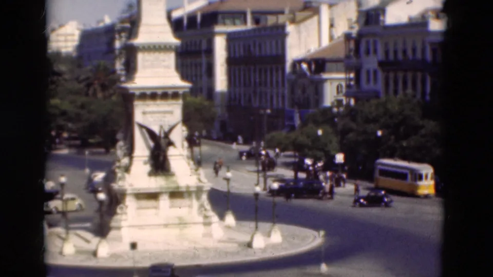 1975 Portugal home-movie frame showing a central monument with winged sculpture, yellow trams, cars, pedestrians, and civic buildings.