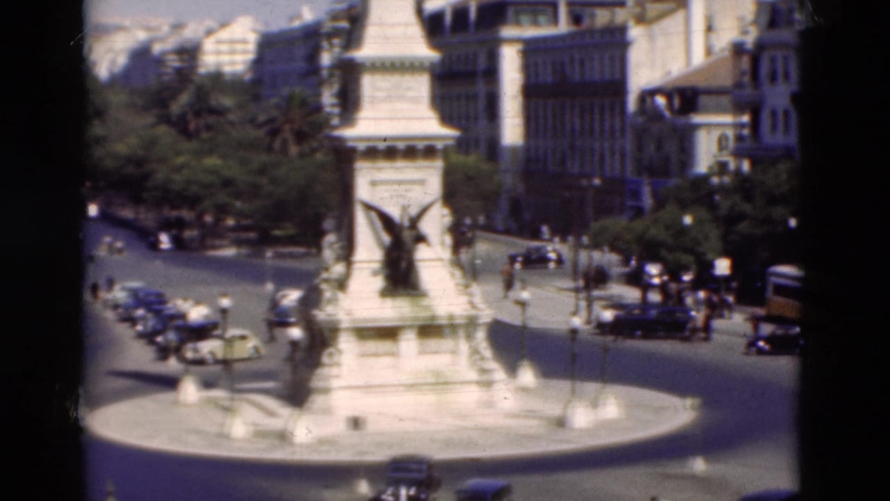 1975 Portugal 8mm frame showing a white monument with winged sculpture, traffic, trees, and surrounding city buildings.