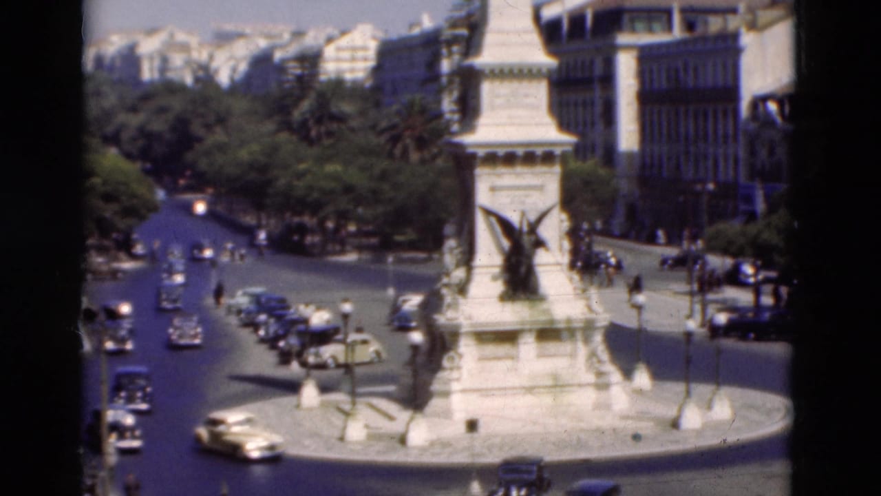 1975 Portugal street scene with classic cars moving around a white civic monument under bright daylight.