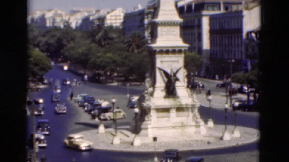 1975 Portugal street scene with classic cars moving around a white civic monument under bright daylight.