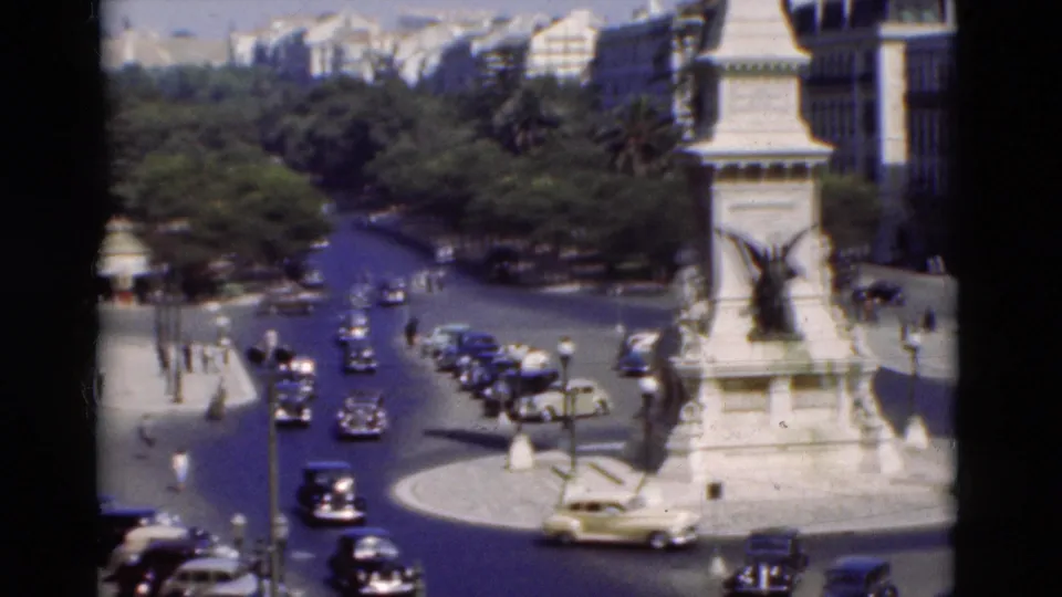 1975 Portugal home-movie frame showing traffic, parked cars, pedestrians, and a tall white monument in a busy square.