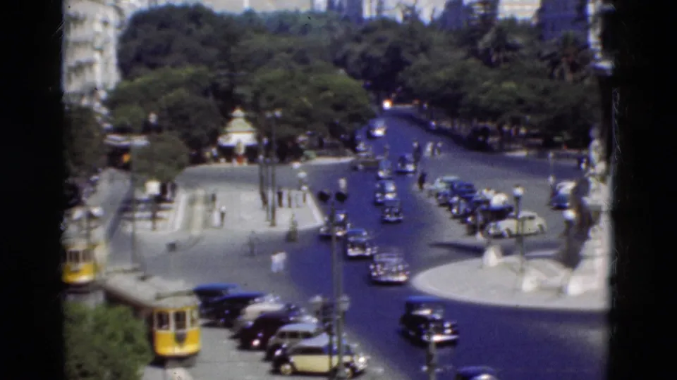 1975 Portugal 8mm view of a yellow trolley, classic cars, trees, pedestrians, and part of a white monument at a city roundabout.