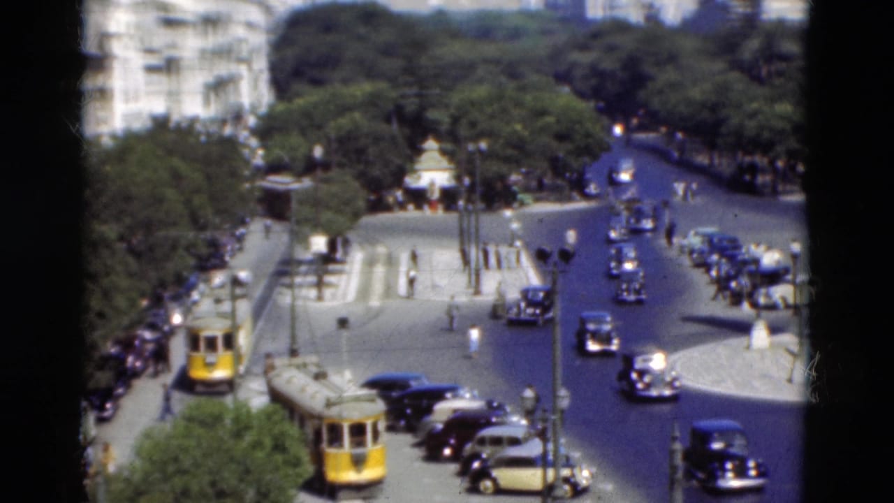 Elevated view of a broad road with vintage cars, yellow transit vehicles, trees, and pedestrians near a coastal district.