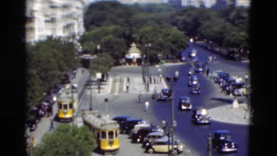 1975 Portugal home-movie frame showing a wide road with classic cars, yellow transit vehicles, trees, and distant pedestrians near the coast.