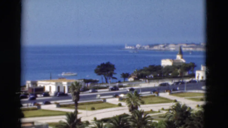 1975 Portugal coastal view with a waterfront road, blue sea, manicured lawns, palm trees, a small boat, and a white church by the shore.