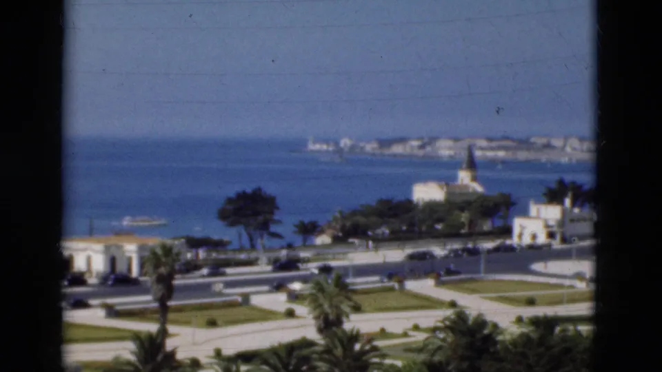 1975 Portugal 8mm frame showing the seaside road, palms, lawns, distant shoreline, small boat, and white church in soft focus.