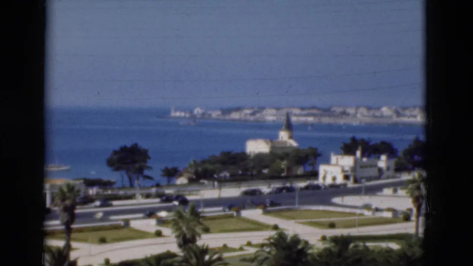 1975 Portugal home-movie frame of a landscaped coastal road, passing cars, blue sea, palm trees, and a white church near the shoreline.