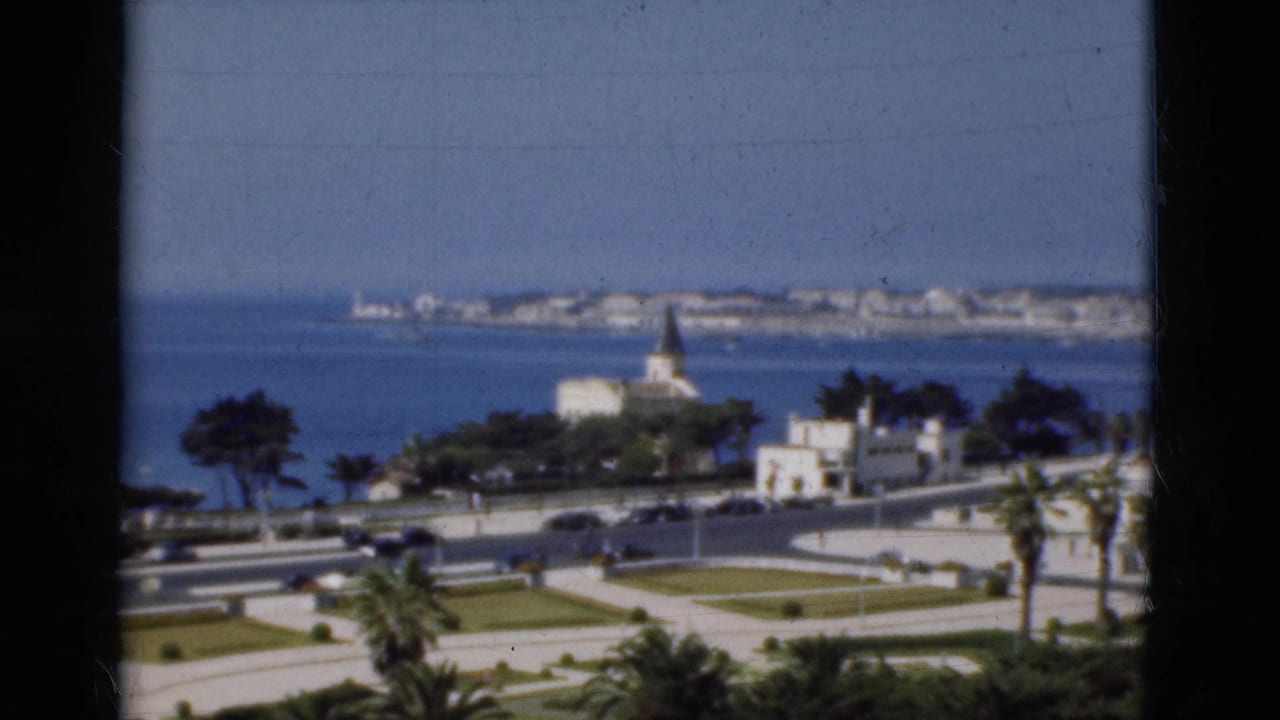 1975 Portugal home-movie frame showing a coastal cityscape with blue sea, palm-lined roads, white buildings, cars, and a pointed steeple.
