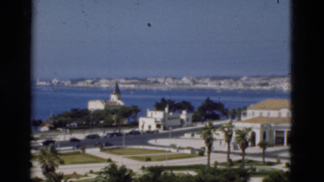 1975 Portugal frame of a waterfront cityscape, with dark film edge framing sea, palm trees, road traffic, white buildings, and a steeple.