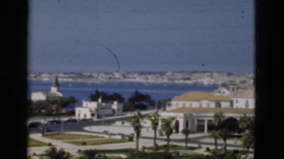 1975 Portugal home-movie frame showing a white waterfront building, palms, curved road, sea, distant town, and visible film marks.