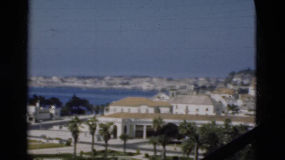 1975 Portugal frame with white seaside buildings, palm trees, roads, blue water, distant shoreline, and dark 8mm frame edge.