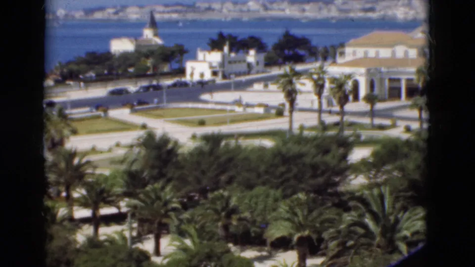 1975 Portugal coastal view with dark window edges, palm-lined grounds, white buildings with pale roofs, and distant city across blue water.